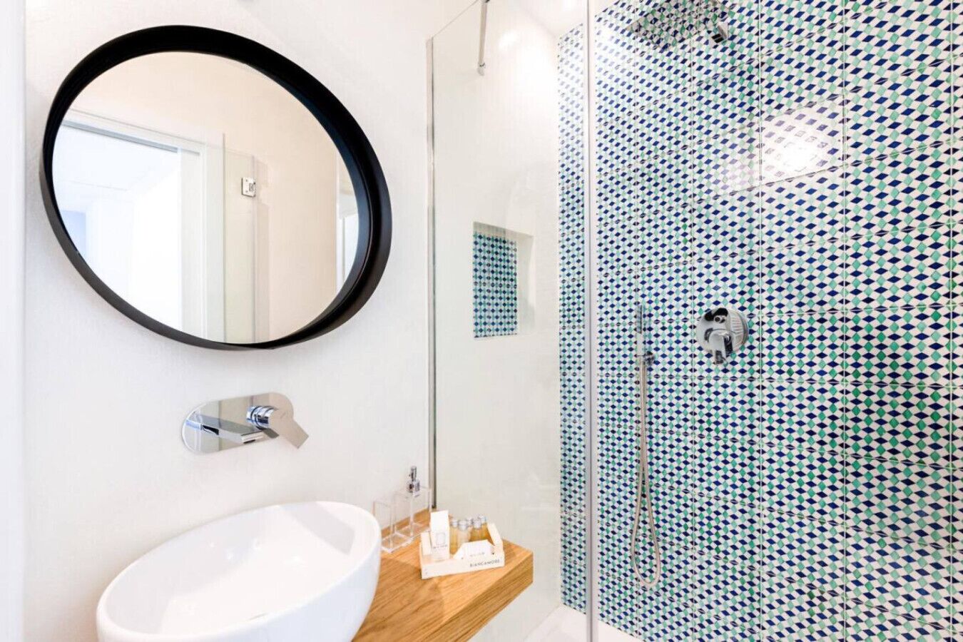 Une salle de bain avec un miroir rond, un lavabo blanc et une douche dont les carreaux sont disposés selon un motif géométrique.