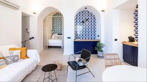 Una habitación de hotel moderna con un sofá blanco, una silla negra, una mesita redonda y un mostrador de recepción con baldosas estampadas.