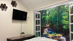 A bedroom with a bed, a TV mounted on the wall, a small table with a plant, and a window with a nature scene view. - 🎰🚶‍♂️ Steps to Caesars 🌊 Riverfront 🍽️ Dining 💦 Hot Tub Add-On 🛏️ Sleeps 9 (Windsor)