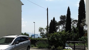 A silver car parked in a driveway with a white building on the left, a metal fence, and tall trees in the background. - Charming and Cozy Apartment Crikvenica (Crikvenica)