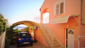 A blue car parked in a narrow driveway leading to a house with a pink facade, a white staircase, and a view of the sea. - Lush and Sunny Apartment With Balcony and Sea View (Crikvenica)
