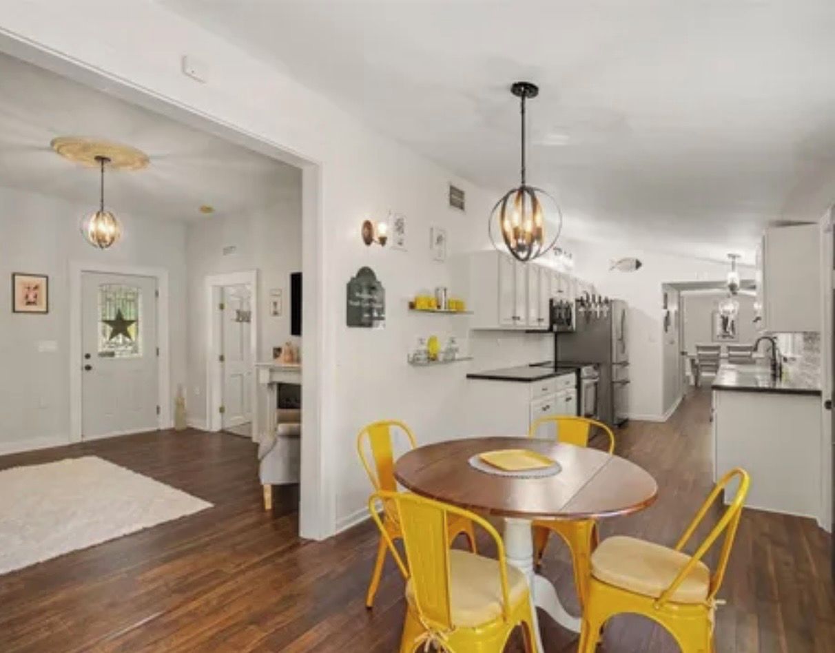 A dining area with a round table and yellow chairs, a kitchen in the background, and a white door with a star decoration.