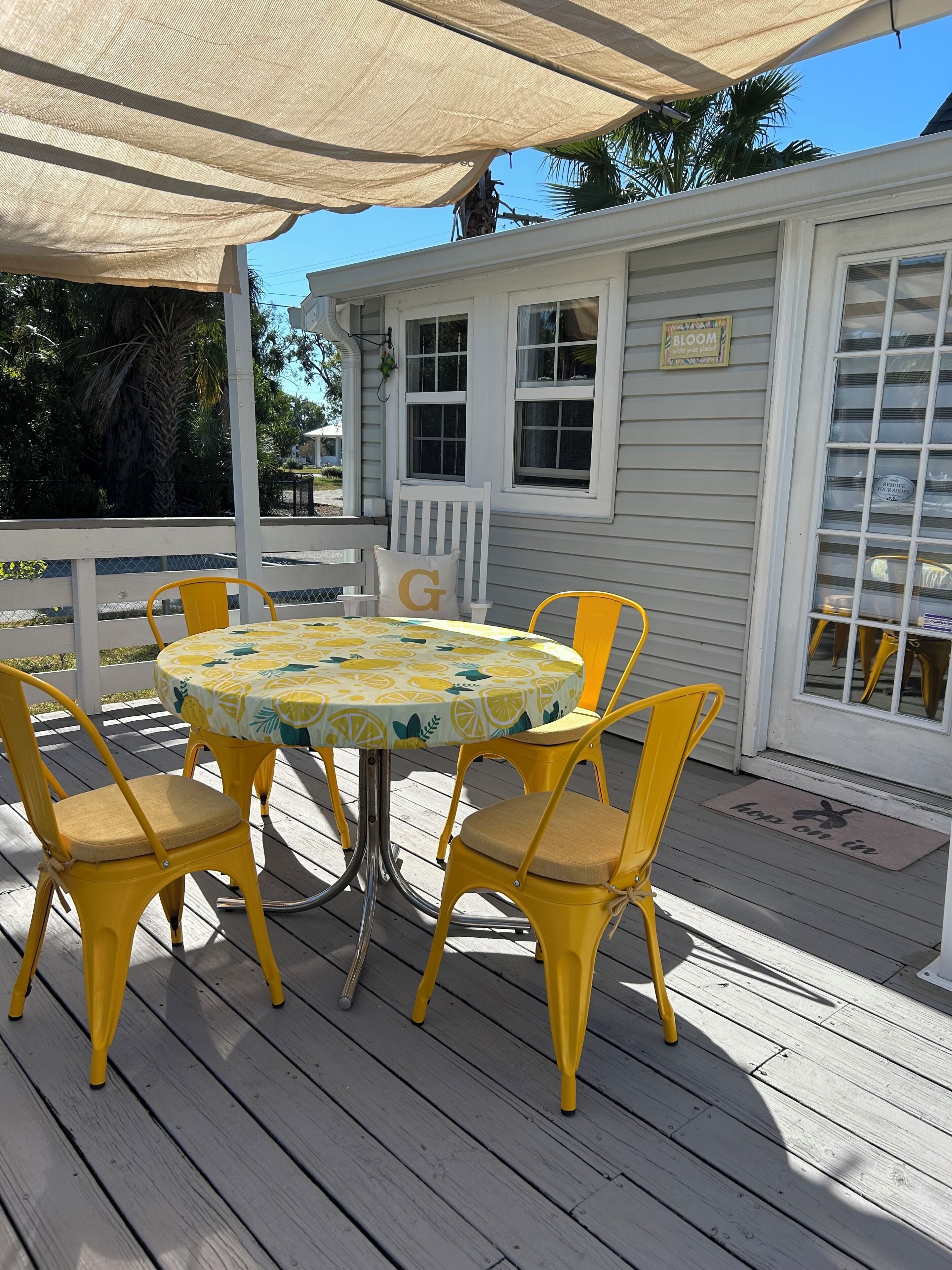 A covered outdoor patio with a round table and yellow chairs, a white building with windows, and a palm tree in the background.