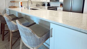 A modern kitchen with white cabinets, a stainless steel refrigerator, a microwave, and a coffee maker. There are two bar stools with woven seats.