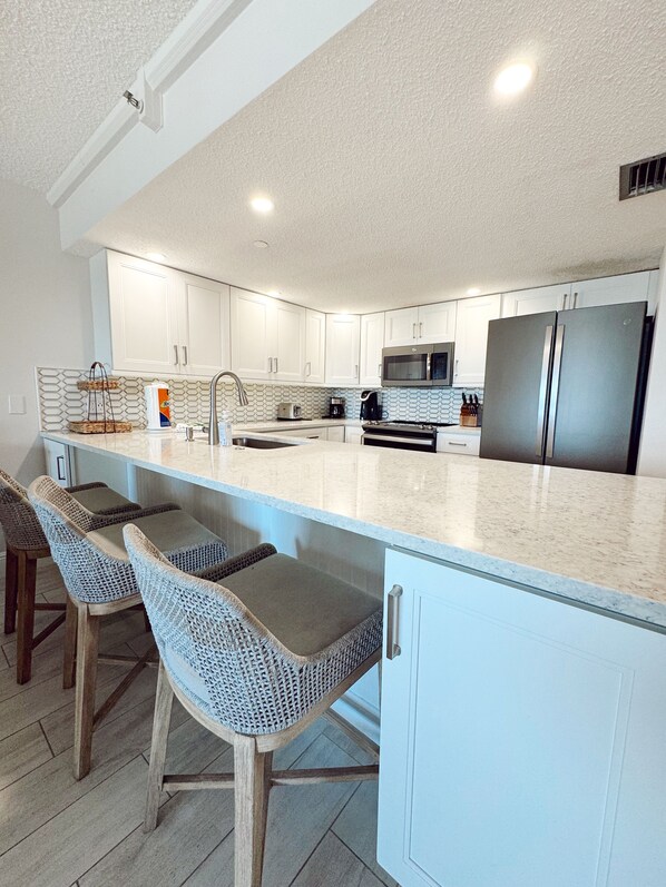 A modern kitchen with white cabinets, a stainless steel refrigerator, a microwave, and a coffee maker. There are two bar stools with woven seats.