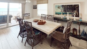 A dining area with a large table, wicker chairs, and a view of the ocean through a sliding glass door.