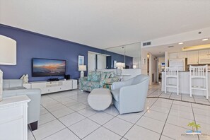 A modern living room with a flat-screen TV, a sofa, a coffee table, and a kitchen area with white cabinets and a refrigerator.