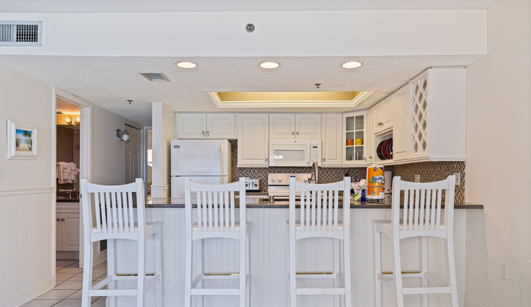 A kitchen with white cabinets, a refrigerator, a microwave, and a sink. There are three white bar stools and a window with blinds.