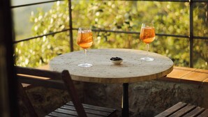 A round table with two glasses of a beverage, a bowl of olives, and a view of green foliage and mountains in the background.