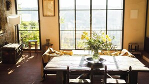 A dining area with a large table, chairs, a vase of flowers, and a view through large windows.