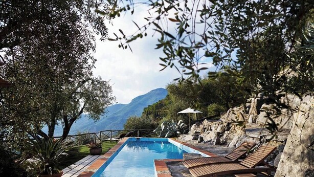 A rectangular pool with a stone border, surrounded by lounge chairs and a rocky landscape with a mountain view.