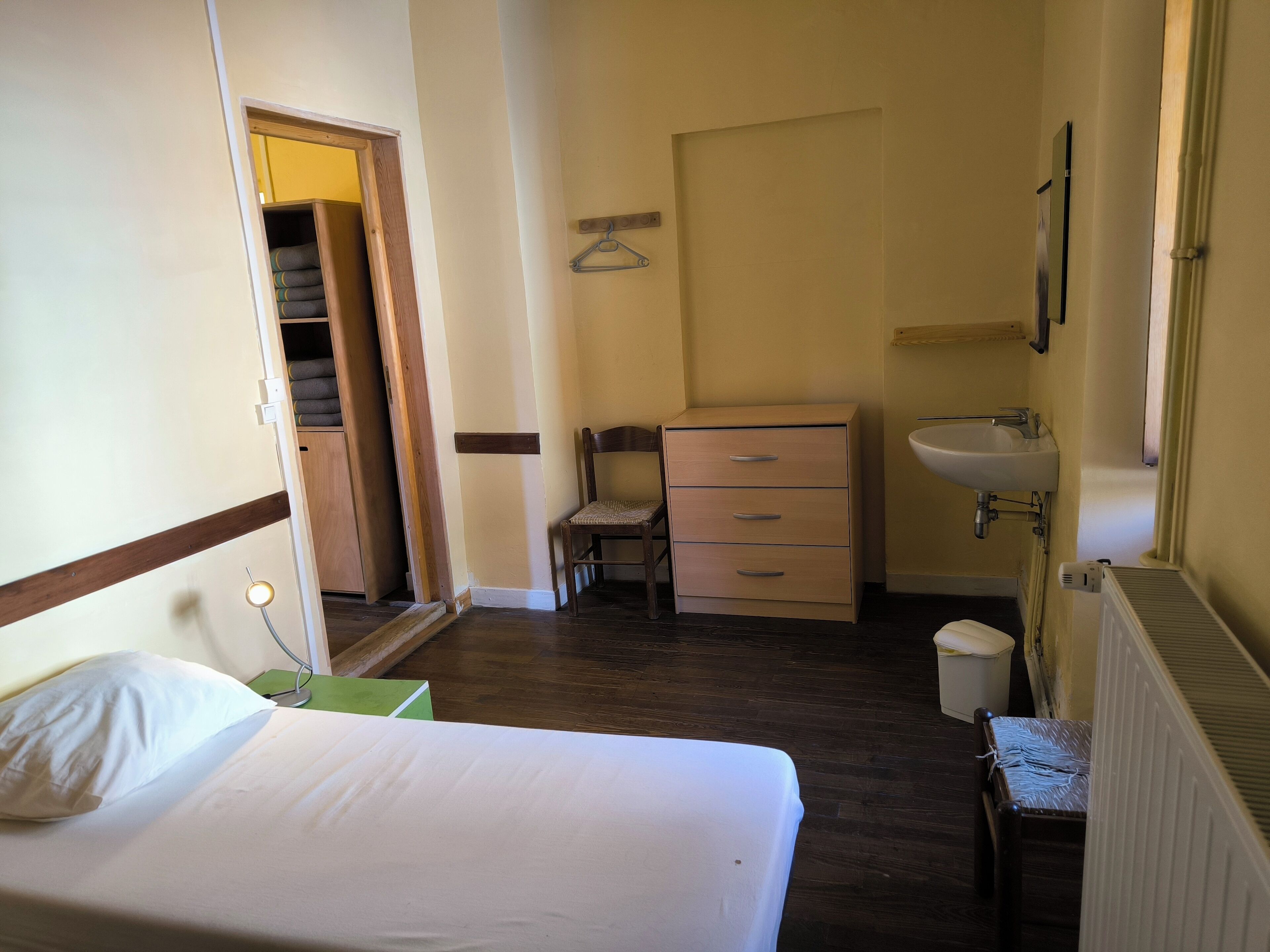 Ein Hotelzimmer mit einem Bett, einem Holzstuhl, einem Schminktisch, einem kleinen Tisch, einer Heizung und einem Badezimmer mit Waschbecken und Spiegel.