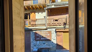 Blick durch ein Fenster auf ein Gebäude mit einer Steinwand und einem hölzernen Balkon. Ein Schild an der Wand zeigt 