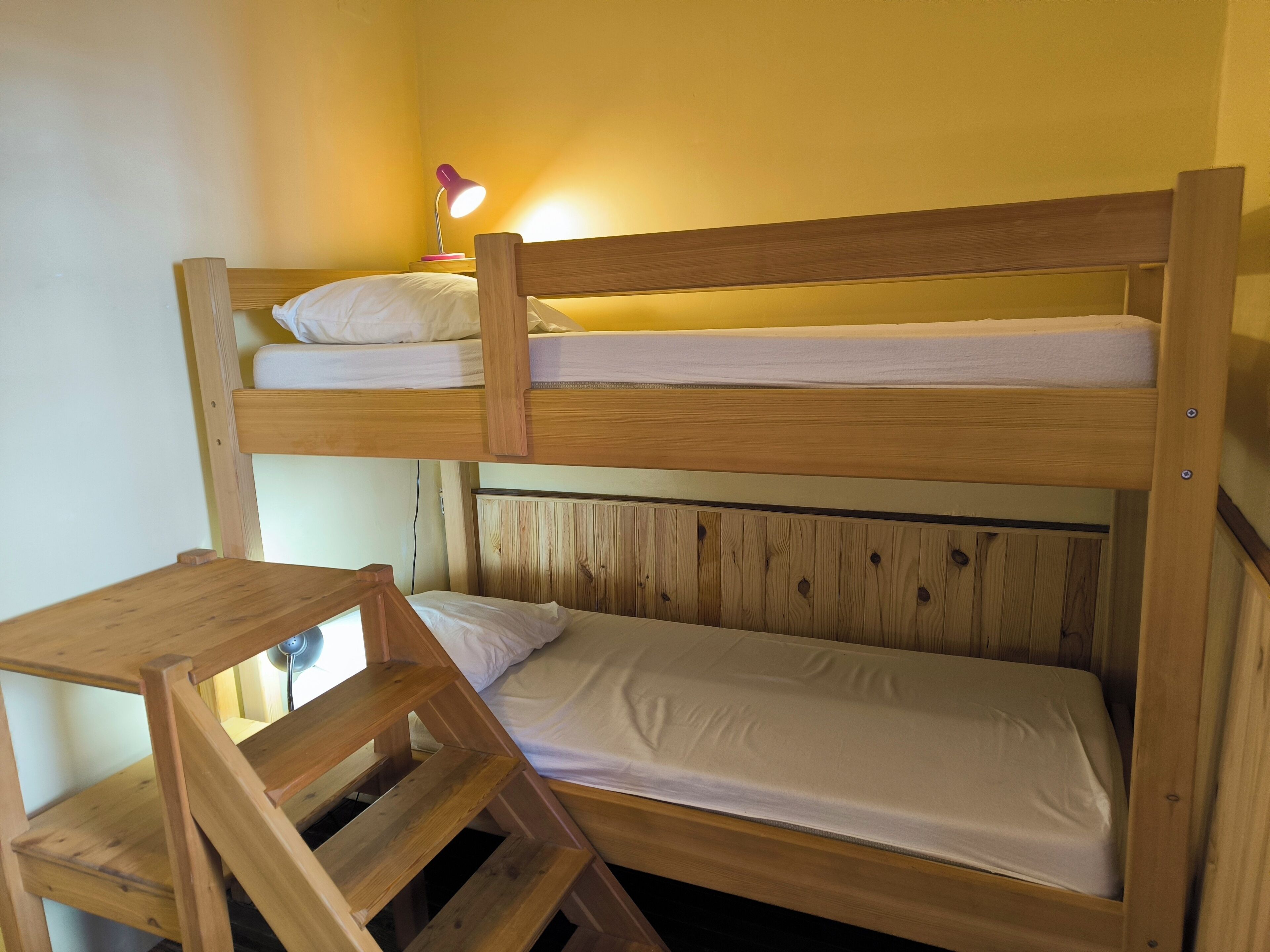 Uma cama de solteiro com estrutura de madeira, uma mesinha e uma escrivaninha.