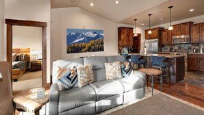 A modern living room with a grey sofa, a dining area with blue chairs, wooden cabinets, and a kitchen with stainless steel appliances. - Moose Mgmt at Black Rock 14151: Hot Tub, Views, Grill, King Beds (Park City)