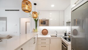A modern kitchen with white cabinetry, a built-in oven, a microwave, and a refrigerator. There are two pendant lights with a woven design. - Seaside Stay With Pool (Estepona)