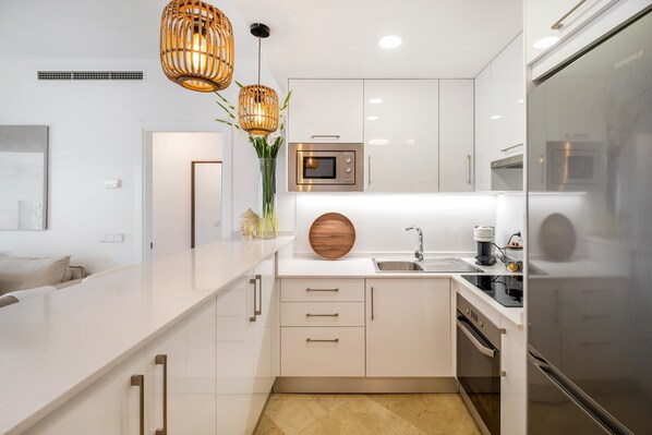 Une cuisine moderne avec des meubles blancs, un four encastré, un four à micro-ondes et un réfrigérateur. Deux suspensions lumineuses au design tressé complètent l’ensemble.