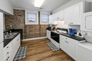 A kitchen with white cabinets, a brick wall, a window, a stove, and a coffee maker. - Lofty Expectations - Gorgeous Studio Loft Downtown (Maryville)
