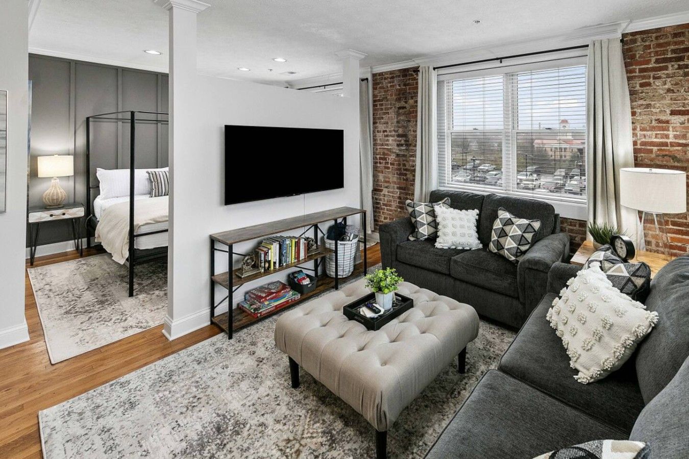 Ein modernes Wohnzimmer mit einem grauen Sofa, einem beigefarbenen Hocker, einem Bücherregal und einem großen Fenster mit Blick auf die Stadt.