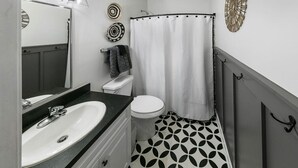Ein Badezimmer mit weißem Waschbecken, Toilette und Duschvorhang. Zwei Wandleuchten, ein Handtuchhalter und dekorative Teller sind vorhanden.