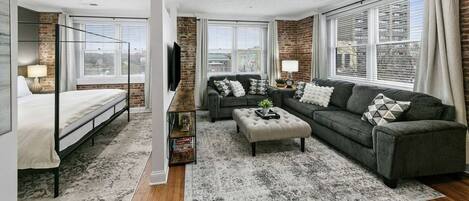 A modern living room with a bed, sofa, and ottoman, a brick accent wall, and large windows.
