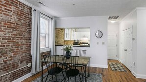 Un comedor con pared de ladrillo, una ventana con persianas, una mesa de comedor con sillas y una cocina al fondo.