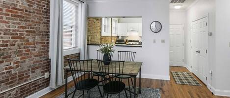 Een eethoek met een bakstenen muur, een raam met zonwering, een eettafel met stoelen en een keuken op de achtergrond.