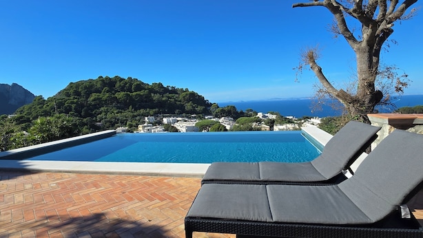 A poolside area with lounge chairs, a clear view of the sea, and a mountainous landscape.