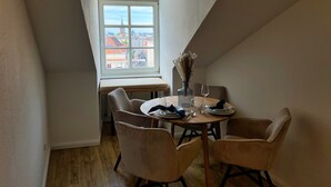 Un comedor con una mesa redonda dispuesta para dos personas, una ventana con vistas a edificios y un banco a lo largo de la pared.