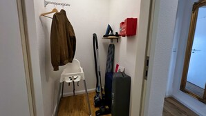 Un pasillo estrecho y pequeño con una pared blanca, piso de madera, una silla blanca, un aspirador, una maleta negra y un perchero con una chaqueta marrón.