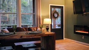 A cozy living room with a fireplace, a television, and a large window overlooking a forest. - Hidden Springs Cottage, Rome, GA, King Loft Bedroom, Private Lake Access (Rome)