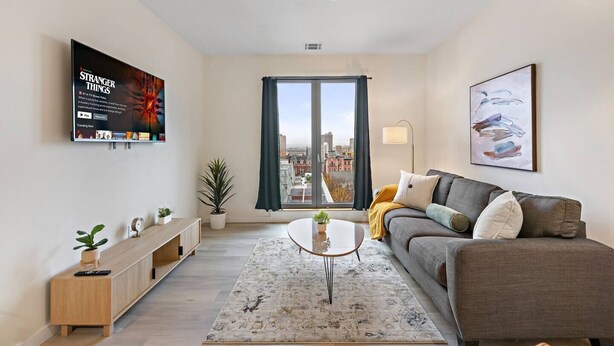 A modern living room with a grey sofa, a coffee table, a TV mounted on the wall, and a city view through the window. - Exceptional 2BR ! Free Parking ! 30 Min to NYC ! (Newark)