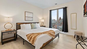 A modern hotel room with a large bed, two bedside tables with lamps, a view of the city through a large window, and a small seating area. - Exceptional 2BR ! Free Parking ! 30 Min to NYC ! (Newark)