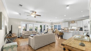 A spacious living area with a sofa, coffee table, and a dining table with chairs. There is a kitchen area with white cabinets and a ceiling fan.
