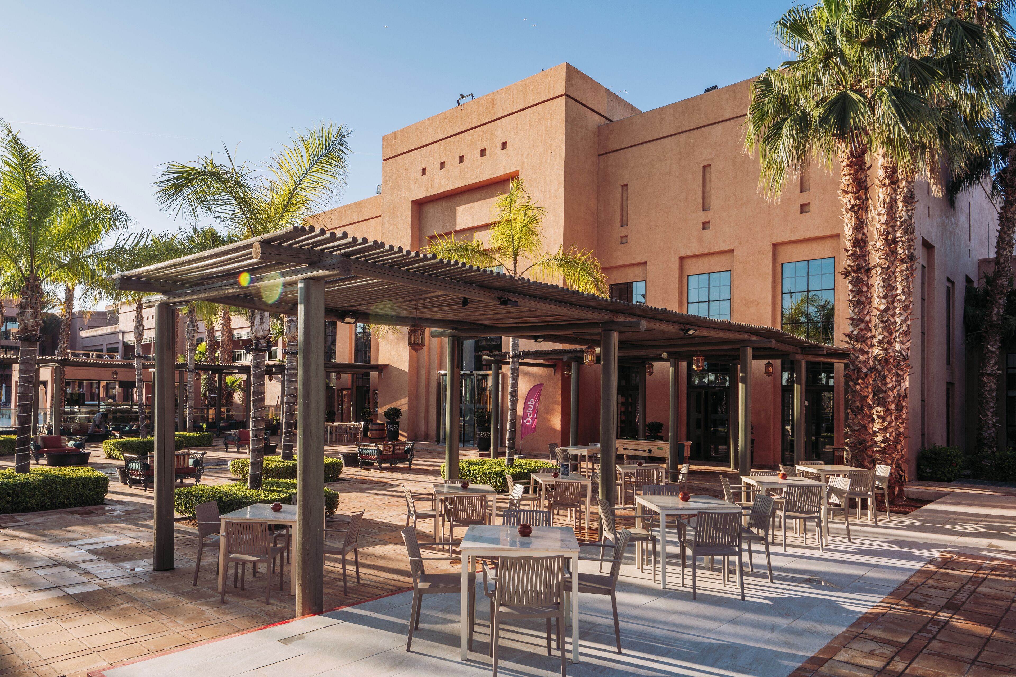 An outdoor patio area with tables and chairs, surrounded by palm trees and a building with large windows.