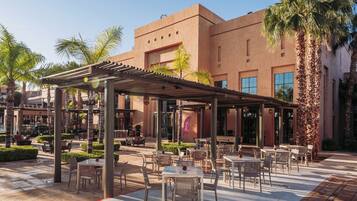 An outdoor patio area with tables and chairs, surrounded by palm trees and a building with large windows.