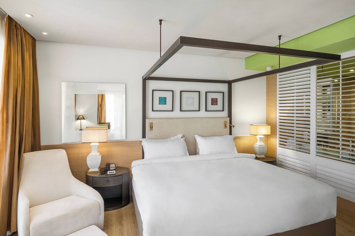 Une chambre d’hôtel avec un grand lit, une table de chevet avec une lampe, une chaise, un miroir et une fenêtre avec des stores.