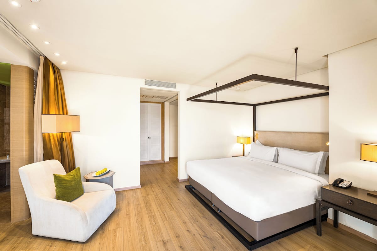 Une chambre d’hôtel moderne dotée d’un grand lit, d’un coin salon avec un fauteuil et une table, ainsi que d’un bureau séparé avec une lampe.