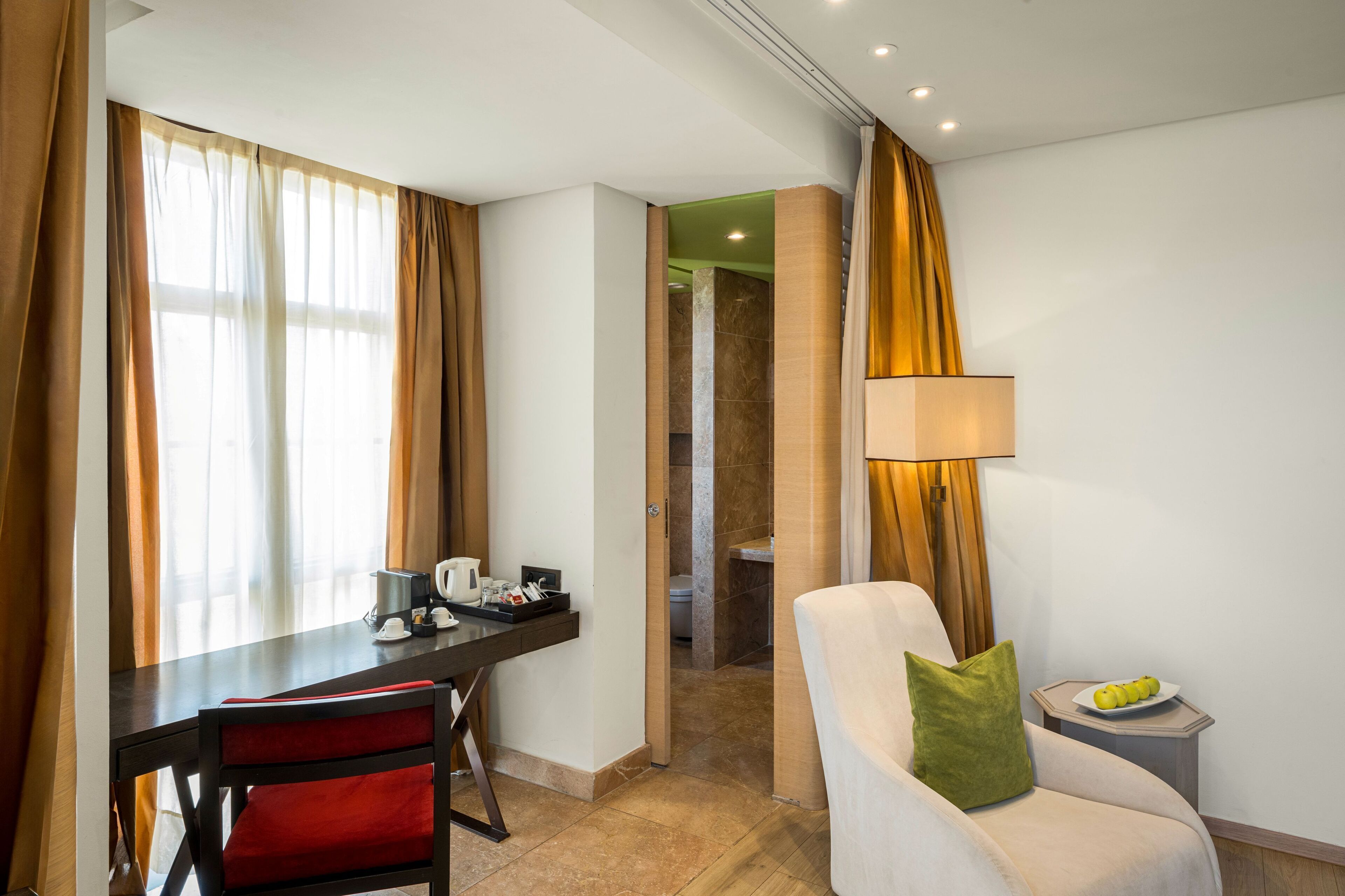 Habitació d'hotel amb zona de menjador, una cadira amb coixí, una làmpada i una taula petita amb fruita.