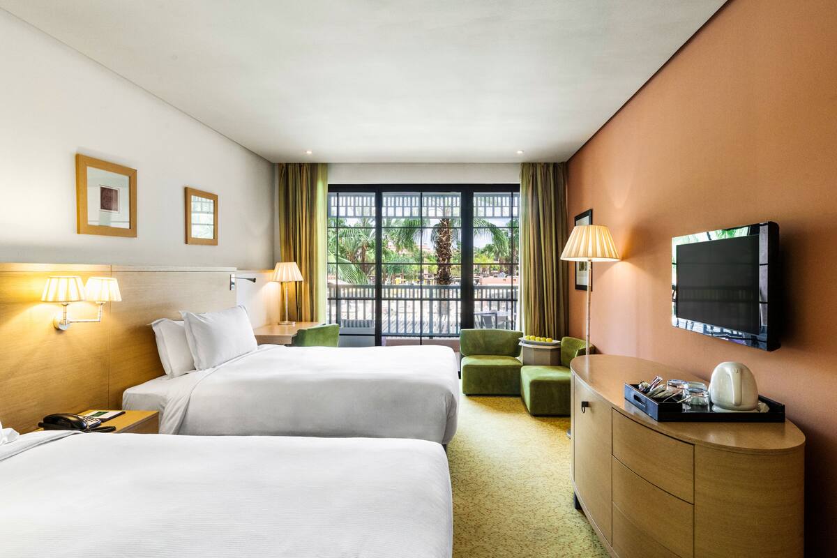 Une chambre d’hôtel avec deux lits, un bureau, une télévision et une vue sur les palmiers, visible à travers de grandes fenêtres.