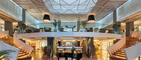 A spacious lobby with a grand piano, a central staircase, and a high ceiling with a geometric pattern.