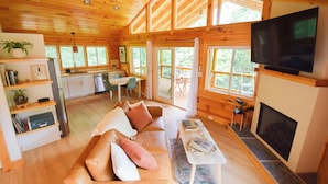 A spacious cabin with a wooden interior, a dining area, a living room with a sofa and fireplace, and large windows offering views of the outdoors. - Cabin, Hot Tub, Fire Pit, 4 miles to Downtown AVL (Asheville)