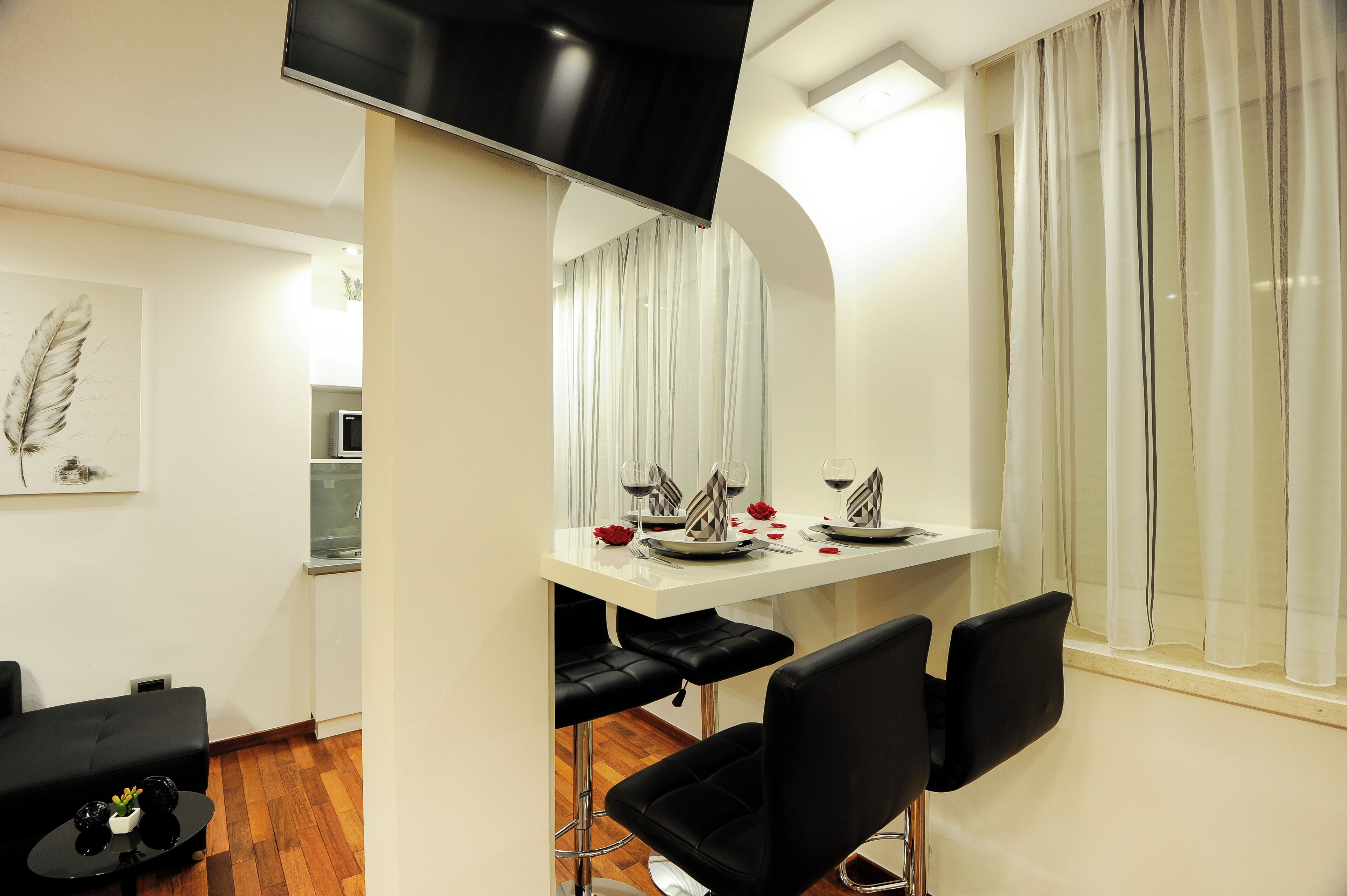 Uma sala de jantar com uma mesa branca posta para quatro pessoas, cadeiras pretas e uma TV montada acima.