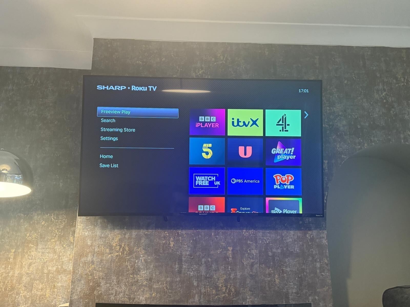 A Sharp Roku TV mounted on a textured wall with a menu interface.