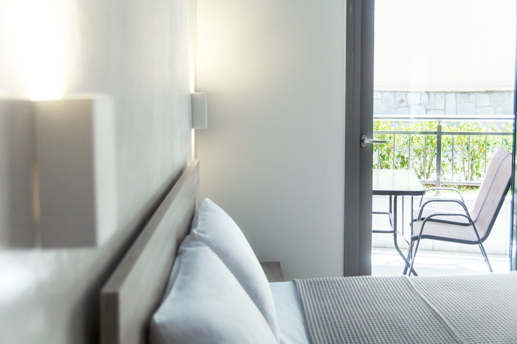 Ein Hotelzimmer mit einem Bett, einem Nachttisch, einer Wandleuchte und Blick auf einen Außenbalkon mit Stühlen und Grünpflanzen.
