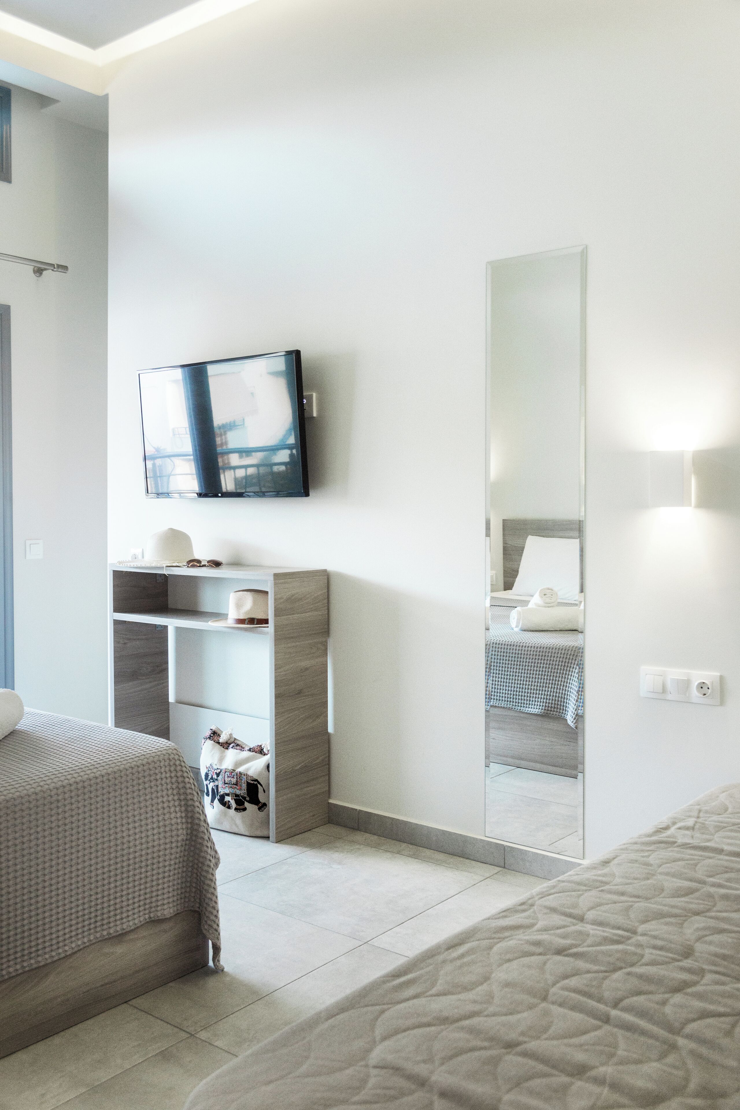 A modern hotel room with a large mirror, a flat-screen TV, a bed with a quilted cover, and a bedside table with a lamp.
