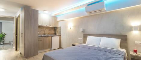 A modern hotel room with a large bed, a bedside table with a lamp, a small kitchenette, and an air conditioning unit.
