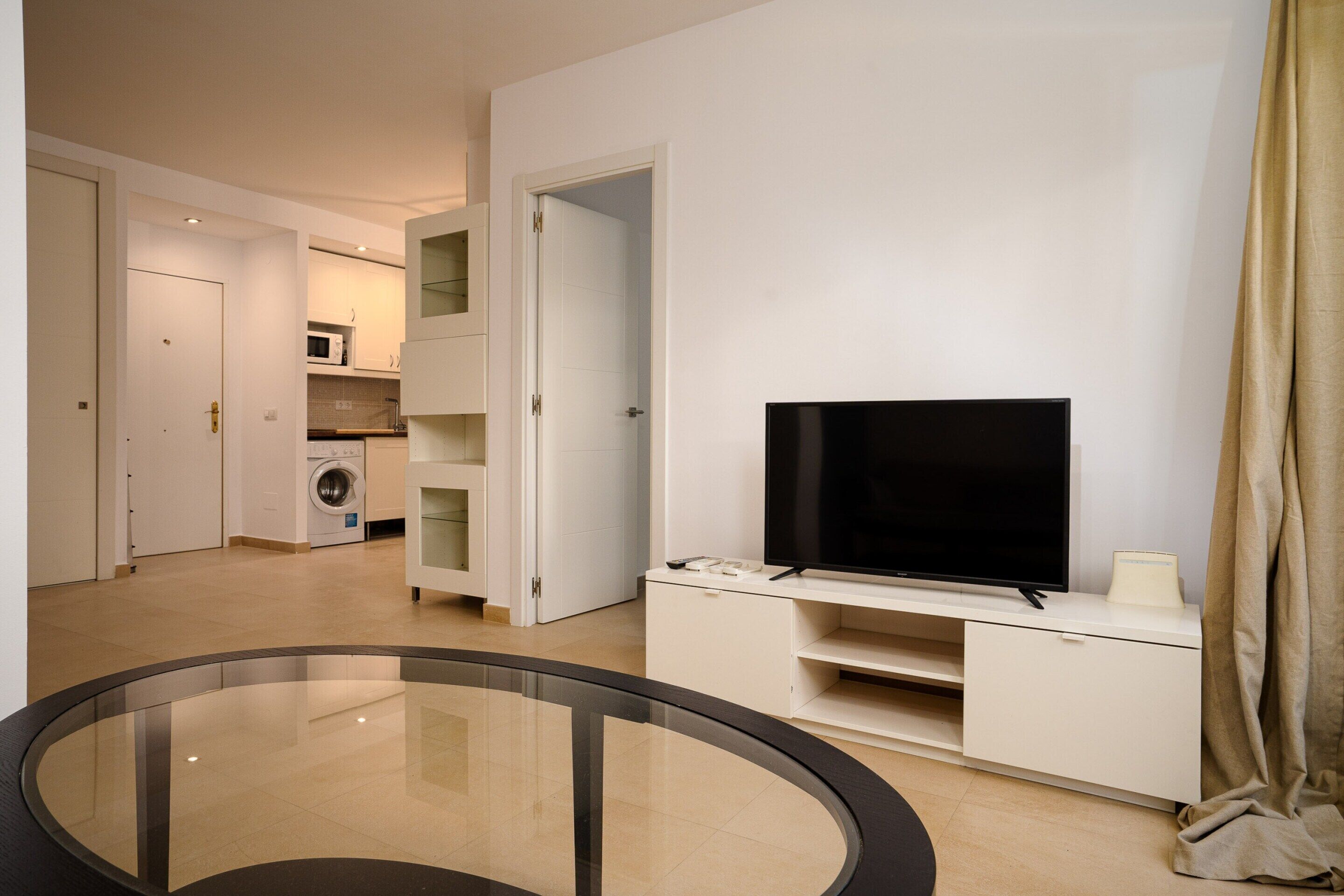 Un salon moderne avec un téléviseur à écran plat posé sur un meuble blanc, une table basse en verre et un coin buanderie à l’arrière-plan.