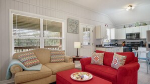 A living room with a beige sofa and a red sofa, both with patterned cushions. There is a small table with a plate of food in front of the red sofa. A kitchen is visible in the background with white cabinets and modern appliances. - Oak St Oasis - Minutes from Downtown and the beach! (Downtown St. Augustine)