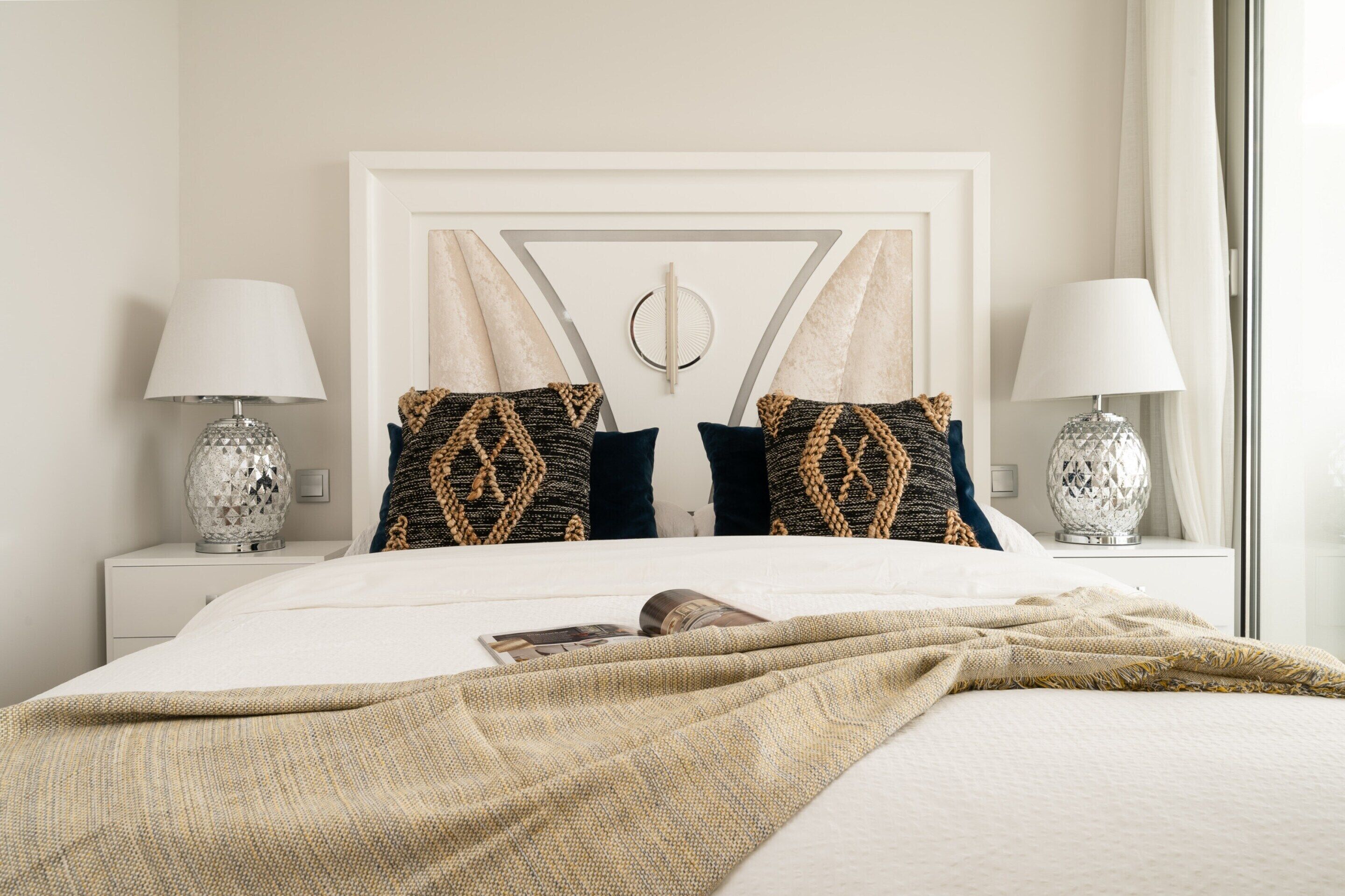 A neatly made bed with a patterned throw blanket, two decorative pillows, and a bedside lamp on a white nightstand.